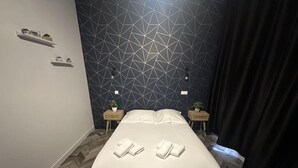 Une chambre moderne avec un mur orné d’un motif géométrique, un lit recouvert de linge de maison blanc, deux tables de chevet agrémentées de plantes et un parquet à motifs en chevrons.