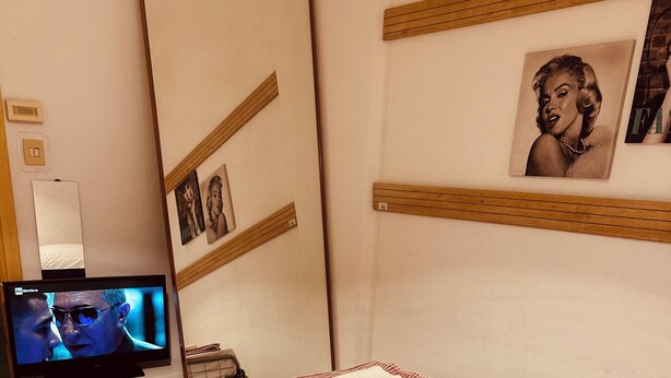 A hotel room with a bed, a television, a wardrobe, and a wall with framed pictures.