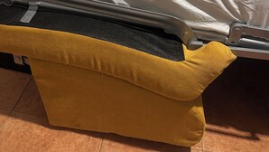 A bed with a white sheet, a pink textured blanket, and a folded white towel. There is a yellow bench with a black cushion in front of the bed.
