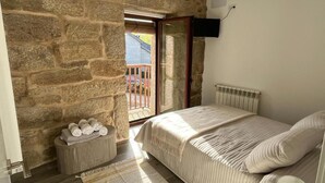 A bedroom with a stone wall, a bed with a white bedspread, a small bedside table with towels, and a window with a view of a balcony.