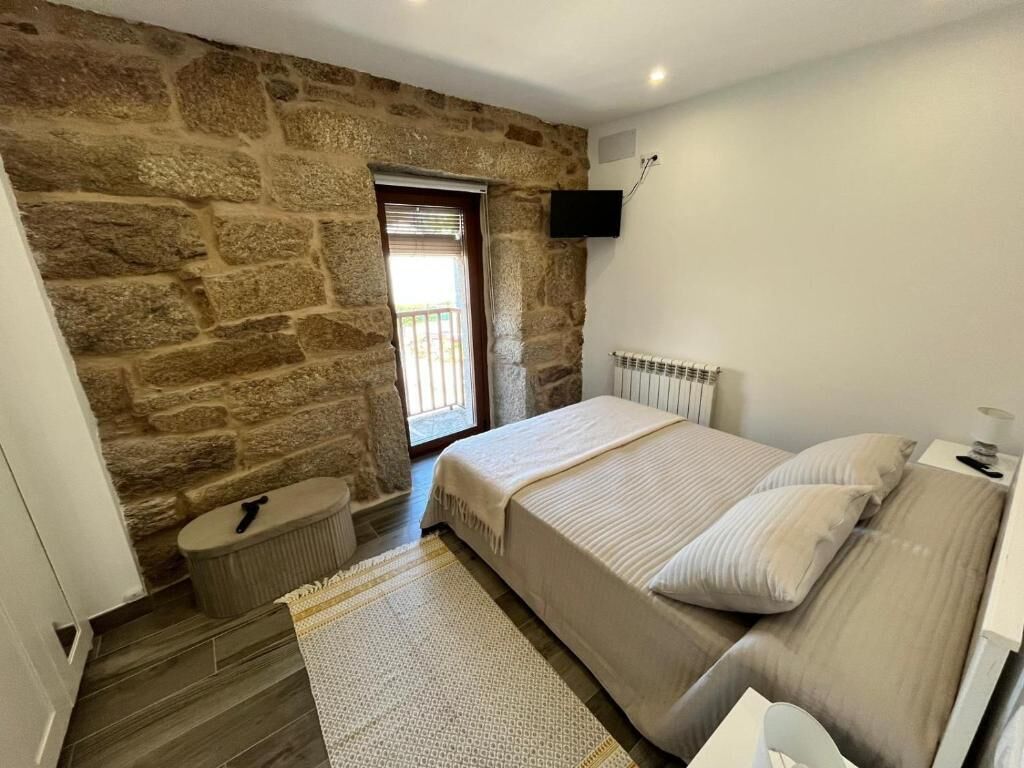 A bedroom with a stone wall, a bed with pillows, a bedside table, a radiator, and a television mounted on the wall.
