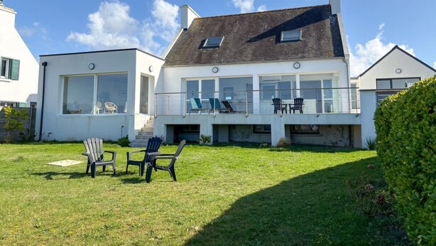Una casa moderna de dues plantes amb un balcó, gespa i mobiliari d'exterior.