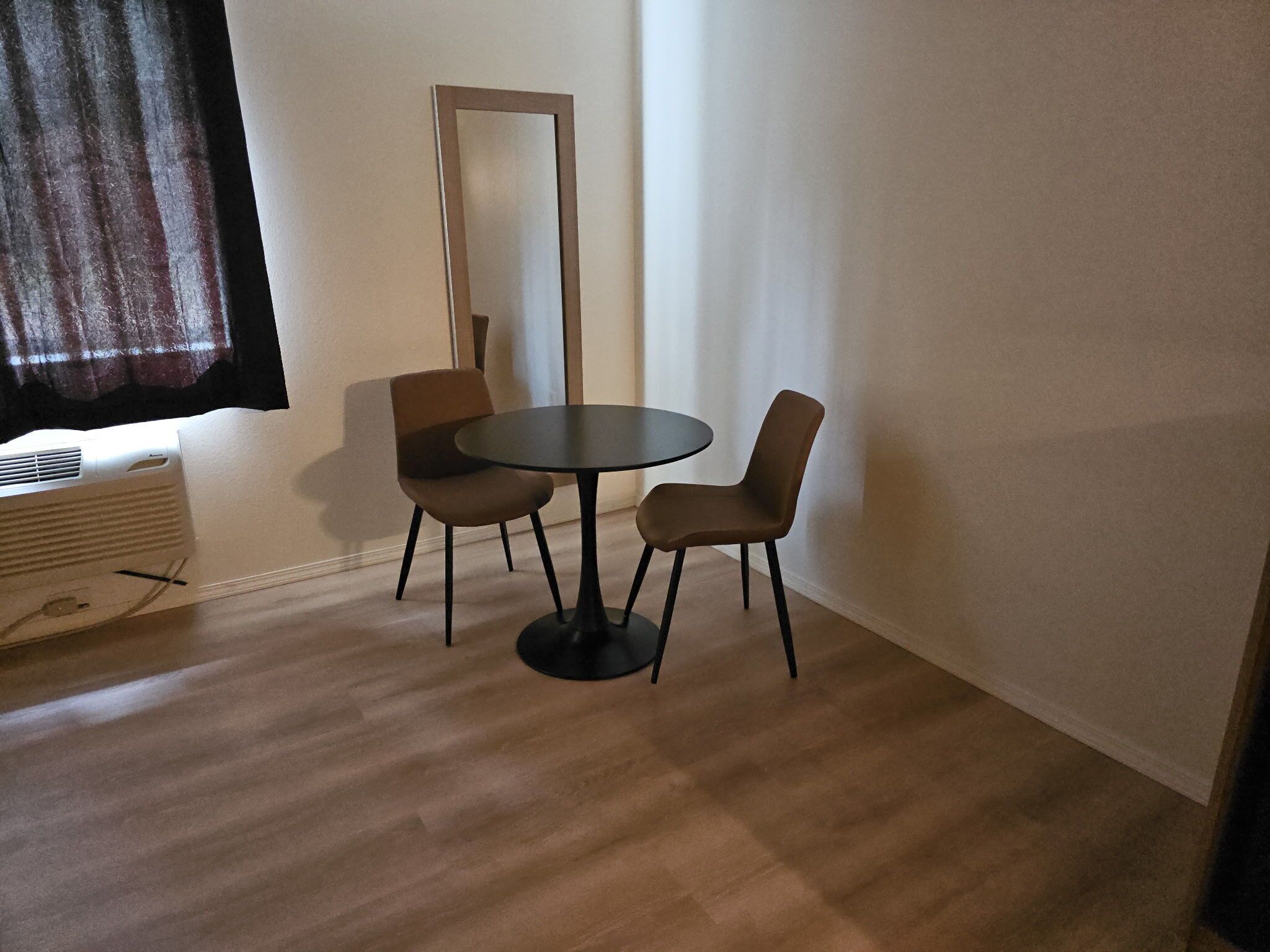 A room with a round table and two chairs, a large mirror on the wall, and a window with curtains.
