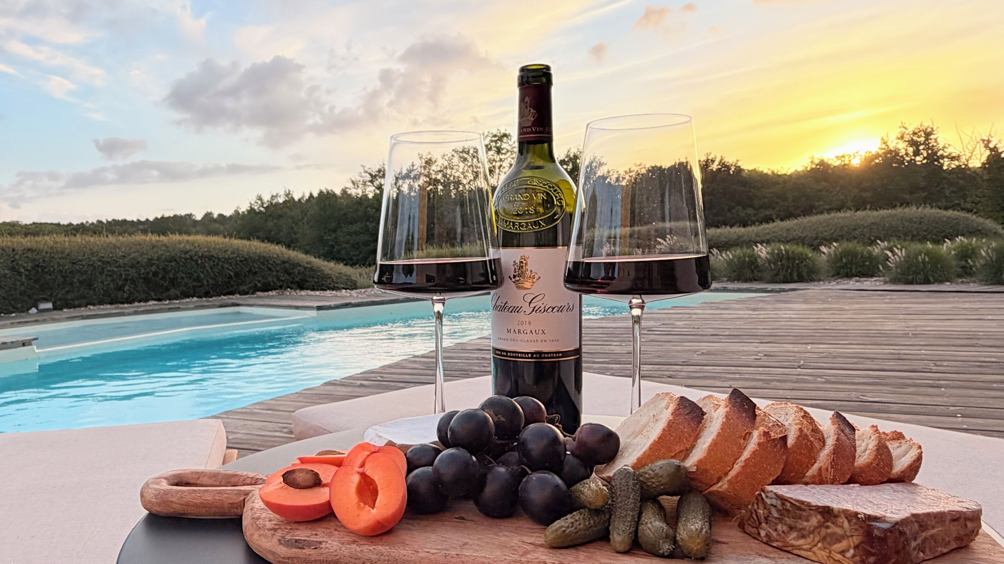Eine Weinflasche, zwei Gläser, ein Holzboden mit Trauben, Brot und Gurken an einem Tisch am Pool.