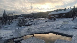 A wooden cabin with a snow-covered roof, surrounded by a snowy landscape and a small pond. - Cottage in the Forest (Lillehammer)