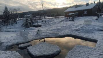 Una cabaña de madera con techo nevado, rodeada por un paisaje invernal y un pequeño estanque.