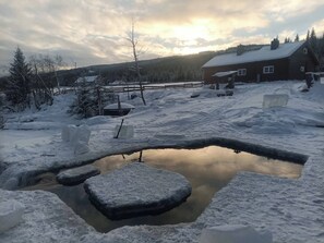 A wooden cabin with a snow-covered roof, surrounded by a snowy landscape and a small pond. - Cottage in the Forest (Lillehammer)