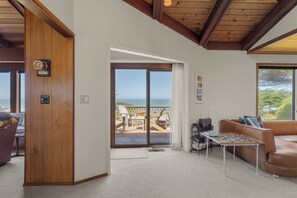 A room with a wooden wall, a sliding glass door leading to a balcony, a brown leather sofa, and a small table with electronic equipment. - Ocean House, by Vinifera Homes (Gualala)