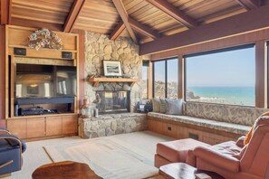 A cozy living room with a stone fireplace, a television, and a large window overlooking a beach. - Ocean House, by Vinifera Homes (Gualala)