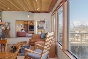 A spacious living area with a large window, a wooden ceiling, and a cozy seating arrangement. - Ocean House, by Vinifera Homes (Gualala)