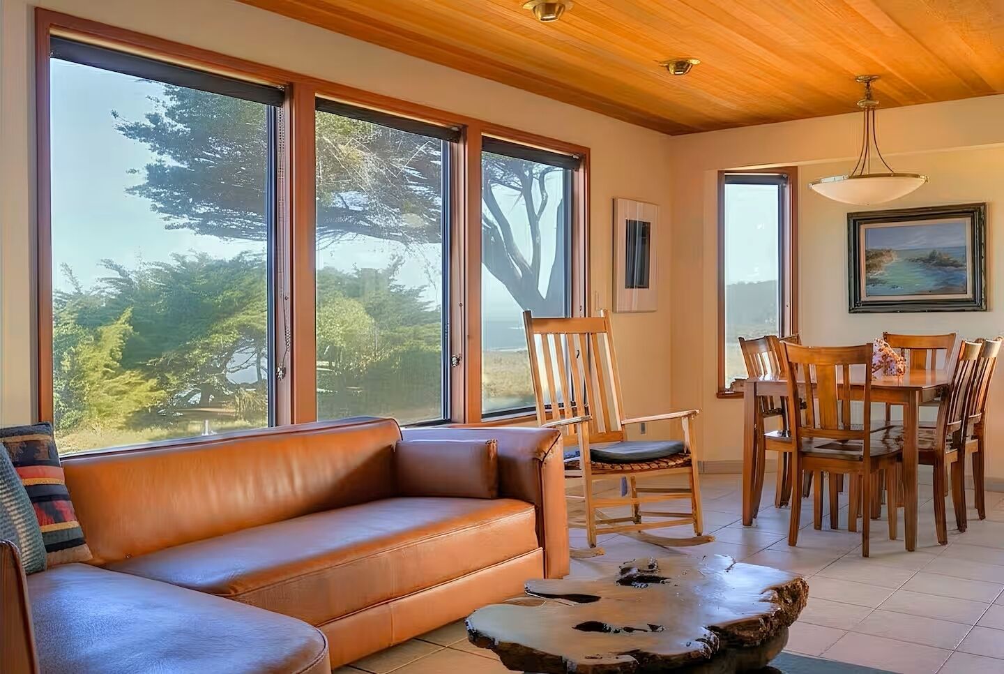 A room with a leather sofa, wooden dining table, chairs, and large windows offering a view of trees.