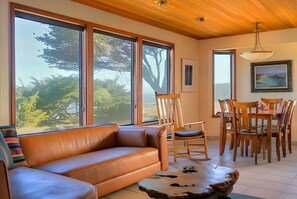 A room with a leather sofa, wooden dining table, chairs, and large windows offering a view of trees. - Ocean House, by Vinifera Homes (Gualala)