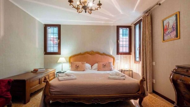 A bedroom with a large bed, a wooden nightstand, a desk, a chair, a chandelier, and a window with curtains. - Grand Canal Palazzo Retreat with Private Canal Views in San Marco (Venezia)