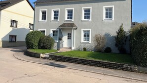 A two-story house with a grey facade, multiple windows, and a small front yard with a hedge.