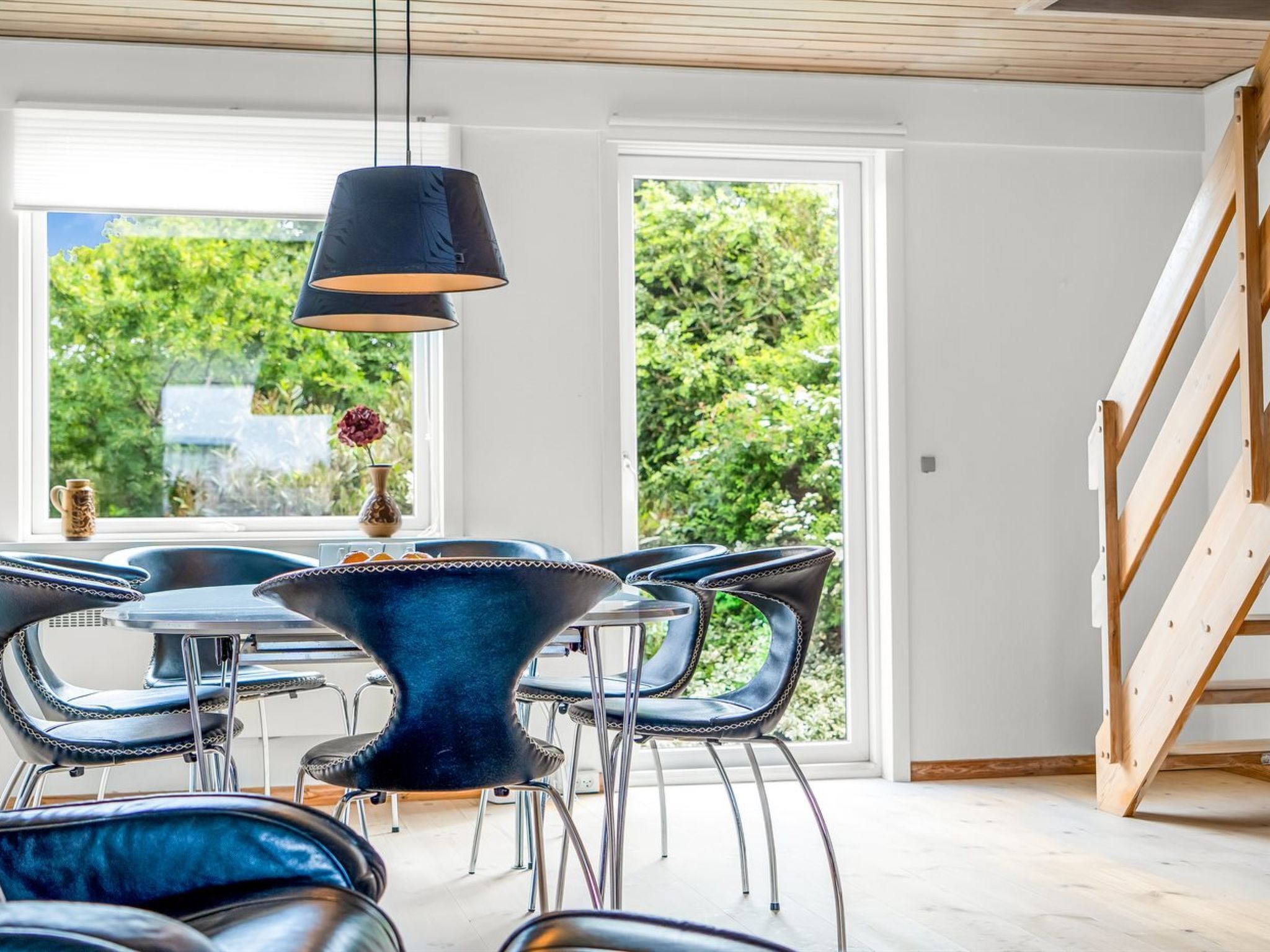 Un espace de repas moderne avec une table ronde et des chaises, un luminaire suspendu et une vue sur la verdure grâce à de grandes fenêtres.