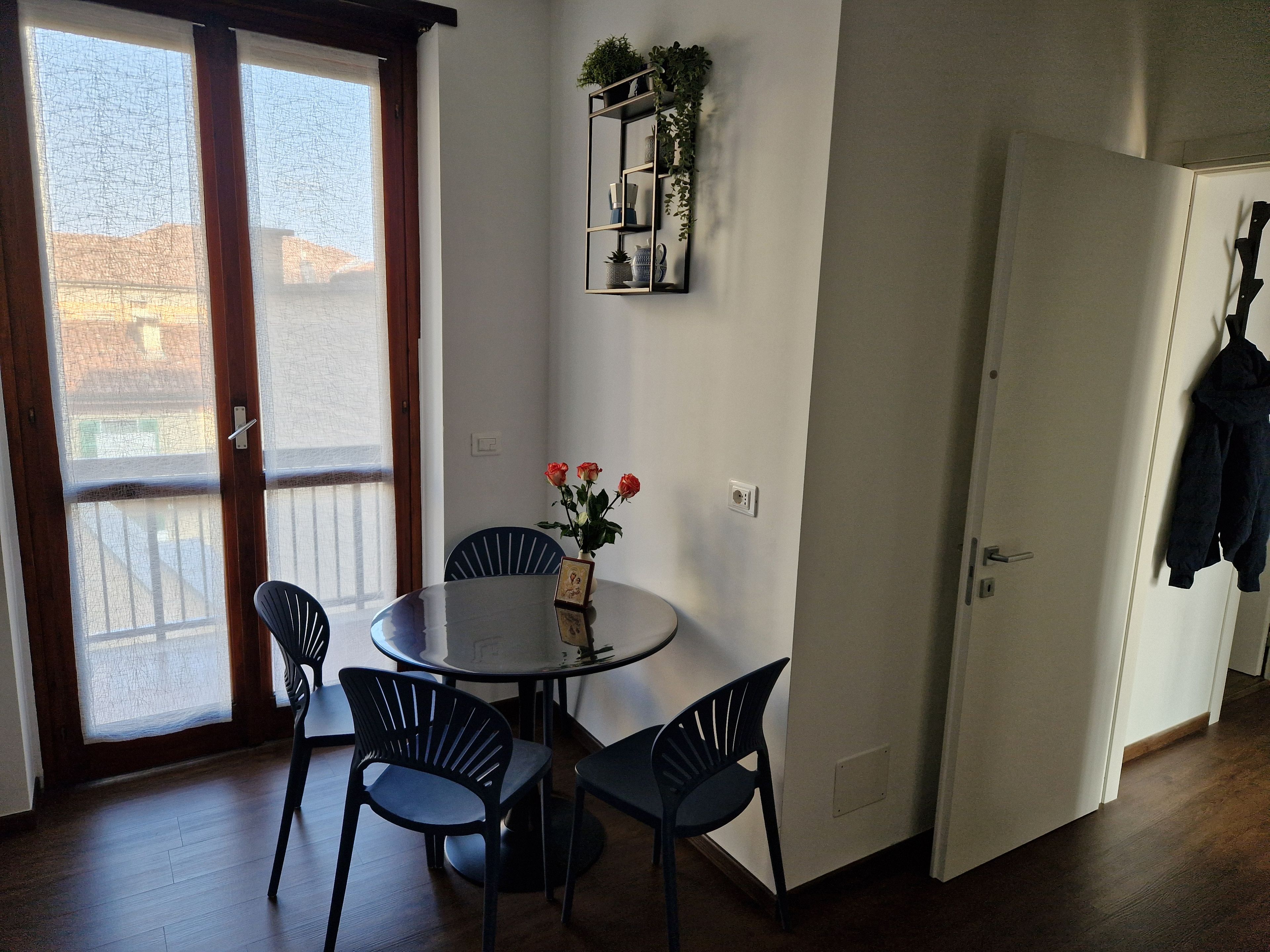 A room with a round table, four chairs, a vase with flowers, a wall-mounted shelf with plants, and a door with a coat hanging.