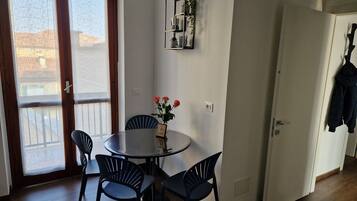 A room with a round table, four chairs, a vase with flowers, a wall-mounted shelf with plants, and a door with a coat hanging.
