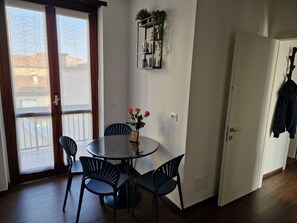 A room with a round table, four chairs, a vase with flowers, a wall-mounted shelf with plants, and a door with a coat hanging. - Beki's Apartment – In the heart of Alba, Piazza Garibaldi (Alba)