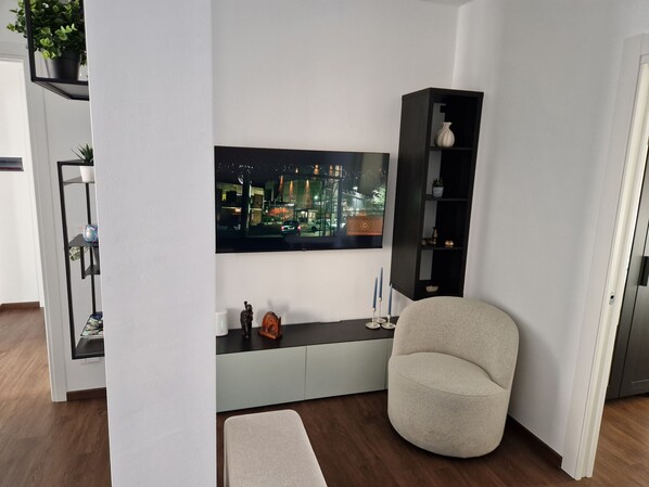 A modern living room with a flat-screen TV mounted on the wall, a grey armchair, a small side table, and a black shelving unit with decorative items.