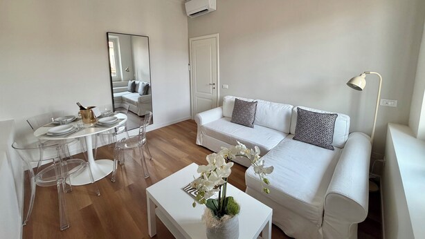Ein modernes Wohnzimmer mit einem weißen Sofa, einem Esstisch mit klaren Stühlen, einem weißen Couchtisch mit einer Blumenvase und einer Stehlampe.