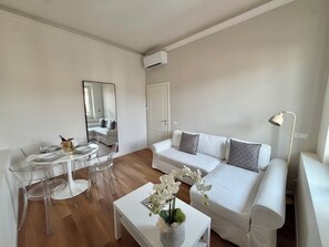 Una sala de estar moderna con un sofá blanco, una mesa de comedor con sillas transparentes, una mesa de centro blanca con un jarrón de flores y una lámpara de pie.