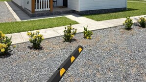A single-story house with a red roof, a porch with a white railing, and a small front yard with gravel and plants. - Leleka's Cabin - E4 (Flowers Bay)