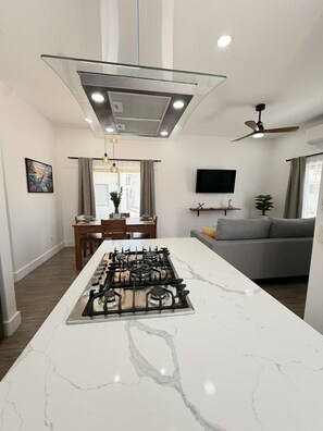 Eine moderne Küche mit Kochinsel, Essbereich mit Tisch und Stühlen, ein graues Sofa und ein an der Wand befestigter Fernseher.