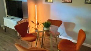Ein Wohnzimmer mit einem runden Glastisch, orangefarbenen Sesseln, einem Fernseher und einer Pflanze.