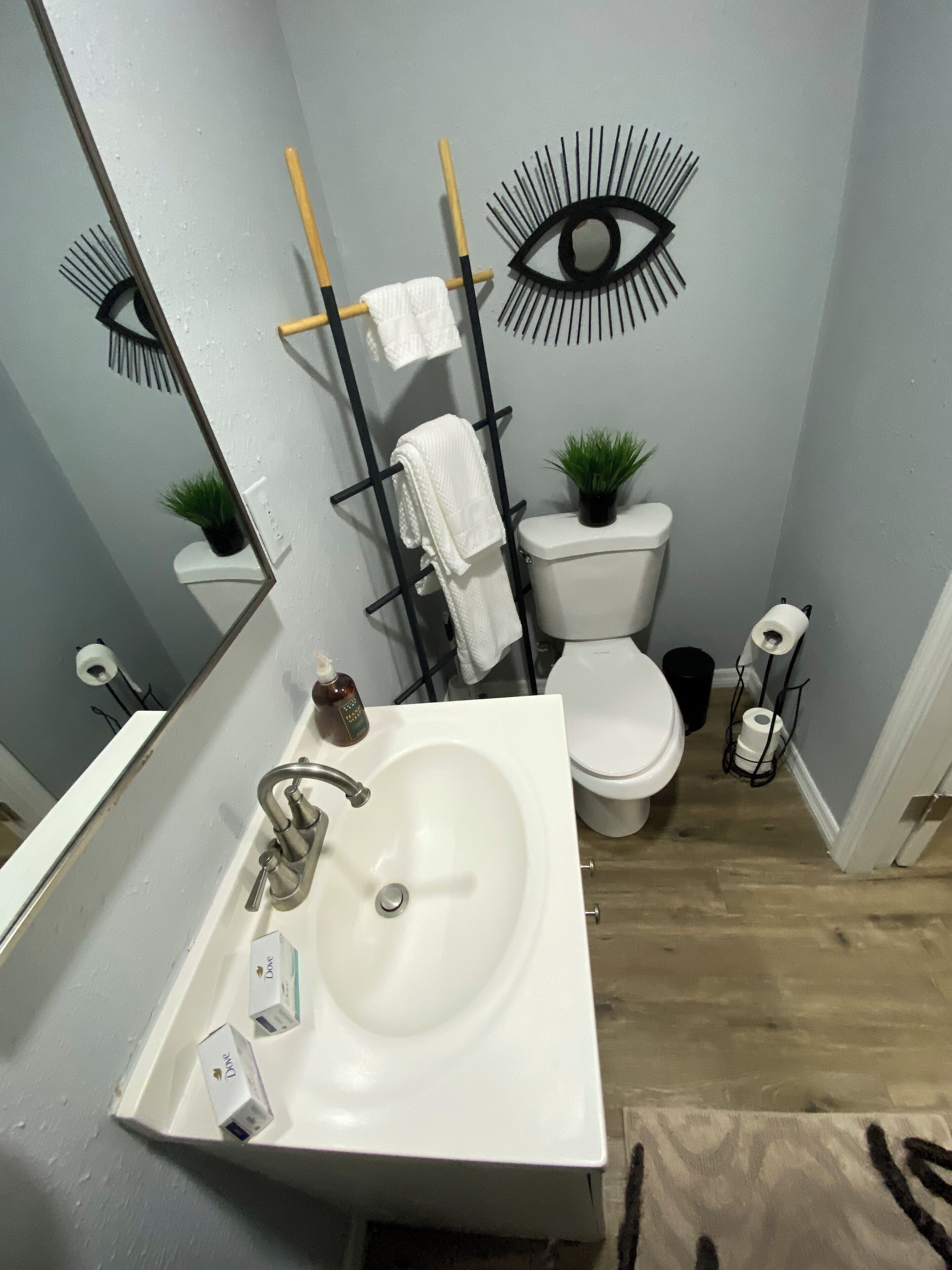 Un bagno con lavandino, water e specchio bianchi. È presente un portasciugamani con un asciugamano, una piccola pianta e un decorativo ornamento a forma di occhio sulla parete.