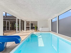 An indoor swimming pool with clear glass walls, a blue slide, and a seating area with chairs and a table. - 5 Star Holiday Home in Glesborg (Glesborg)
