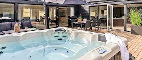 A hot tub on a wooden deck with a covered seating area and a grill in the background.