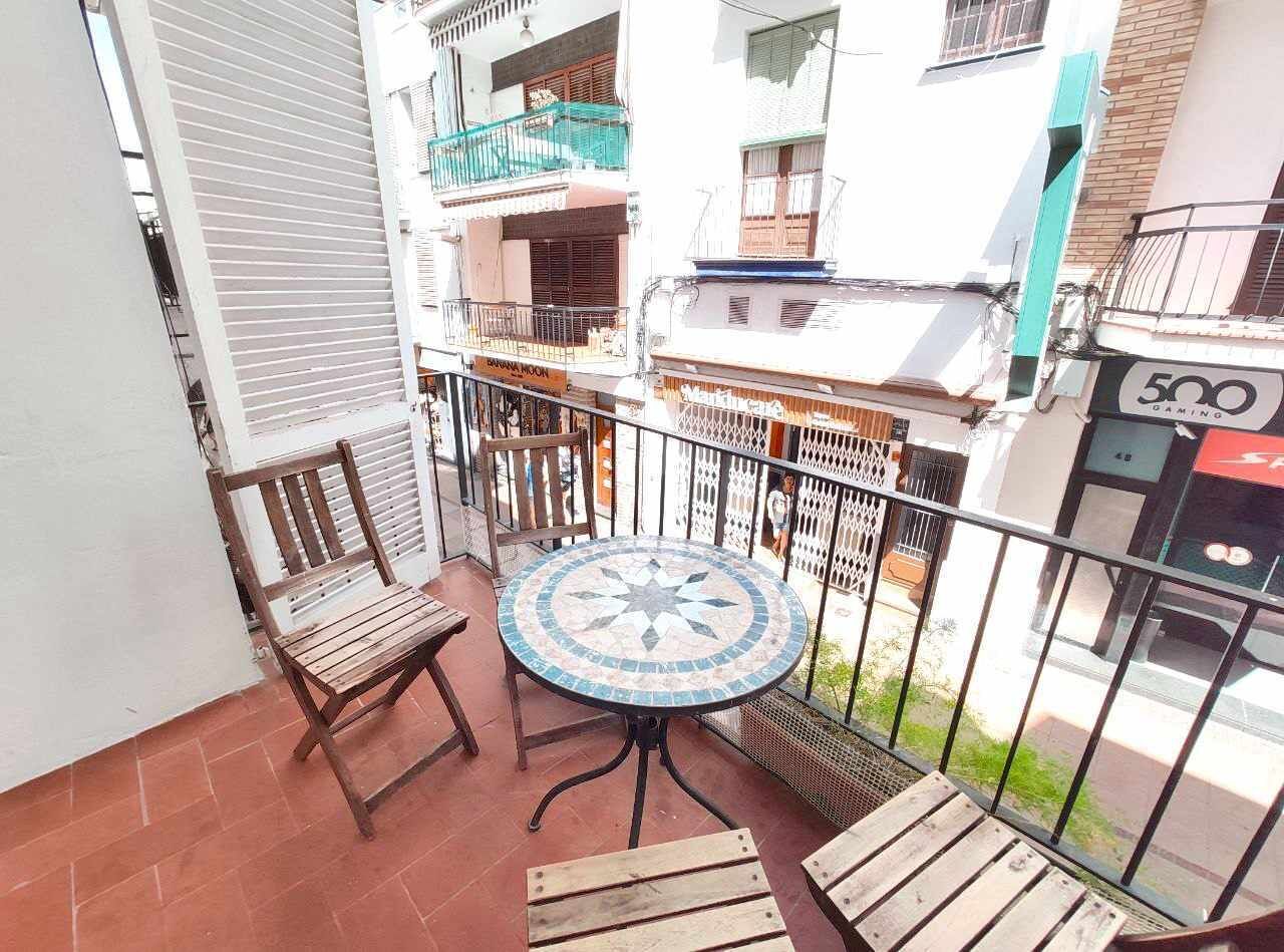 Un balcón con una mesa redonda y dos sillas, suelo de baldosas y vista a un edificio con balcones y un letrero que dice '500 GAMING'.