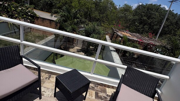 A balcony with wicker furniture, a small table, and a view of a green lawn and trees. - Casa Eterna Primavera  (Playa del Carmen)
