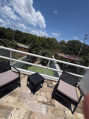 A balcony with wicker furniture, a small table, and a view of a green lawn and trees. - Casa Eterna Primavera (Playa del Carmen)