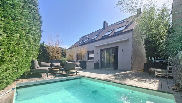 Une piscine avec un espace patio, du mobilier d’extérieur et une maison moderne dotée de grandes fenêtres et d’un toit en pente.