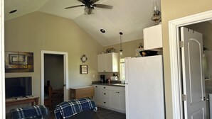 Ein Wohnzimmer mit Deckenventilator, Fernseher, einer Couch und einer Küchenzeile mit Schränken und Kühlschrank.
