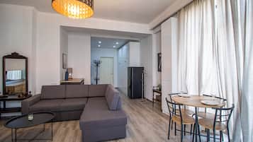 A modern living room with a sectional sofa, a dining table with chairs, a small coffee table, a chandelier, and a view through a doorway.