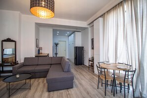 Un salon moderne avec un canapé d’angle, une table à manger avec des chaises, une petite table basse, un lustre et une vue sur l’extérieur, visible à travers une porte.