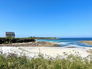 A coastal landscape with a stone building, sandy beach, and clear blue sky.
