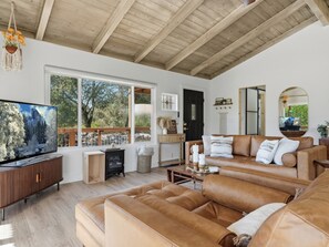 A living room with a television, leather sofa, wooden ceiling, and large windows. - Private. Luxury. River House Retreat at Yosemite. Hot Tub. Outdoor Shower. Views (Coarsegold)