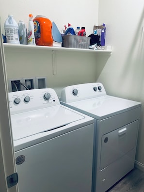 A laundry area with two front-loading washers and a shelf above containing various cleaning supplies.