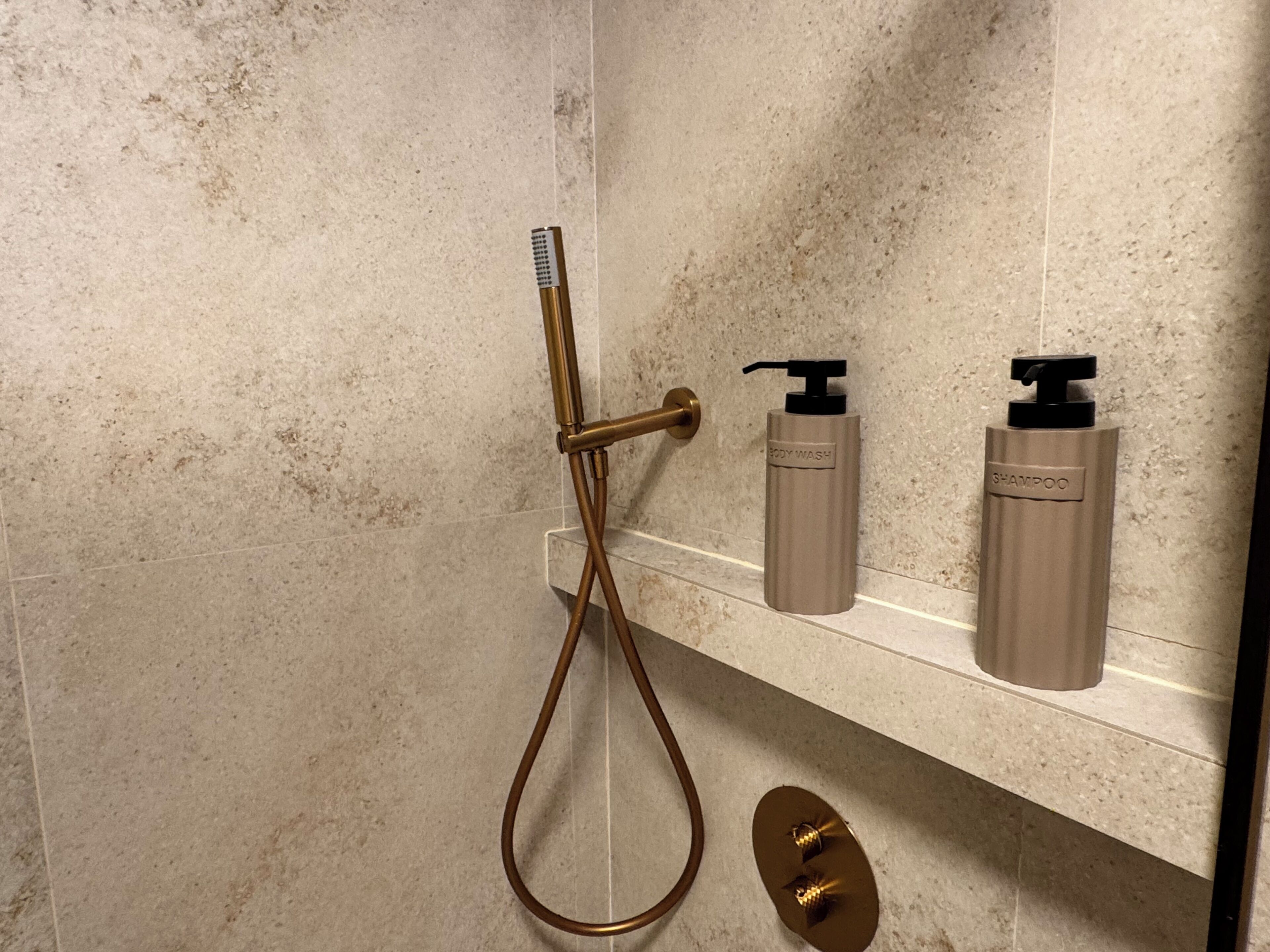 A bathroom with a shower, a hand dryer, and two bottles of shampoo and body wash on a shelf.