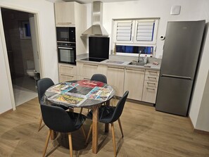 A compact kitchen with a dining table, chairs, a refrigerator, and a window with blinds. - 🎢 Comfortable, Air-Conditioned 2-Room Vacation Rental – Central Alsace – Near Europa-Park (Witternheim)