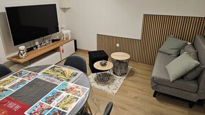 Un salon moderne avec un téléviseur à écran plat, une table basse en bois sur laquelle sont posées des brochures de voyage, un canapé gris et une petite table ronde agrémentée d’un objet décoratif.
