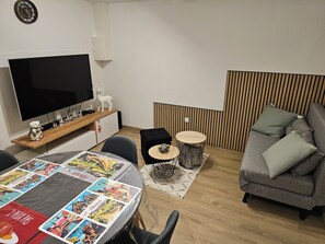 A modern living room with a flat-screen TV, a wooden coffee table with travel brochures, a grey sofa, and a small round table with a decorative item. - 🎢 Comfortable, Air-Conditioned 2-Room Vacation Rental – Central Alsace – Near Europa-Park (Witternheim)