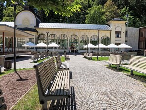 Ein gepflasterter Weg mit Bänken und Sonnenschirmen, ein Gebäude mit Rundbogenfenstern und ein Glockenturm.