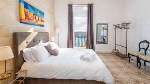A hotel room with a bed, a painting, a window with curtains, a coat rack, and a chair. - Regina Suite: 2-bed, 2 en-suite apartment in Valletta with AC and comfort (Valletta)