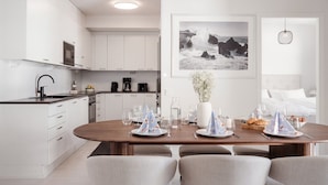 A modern kitchen with white cabinets, a dining area with a wooden table set for four, and a framed photograph above the table. - Large 3BR • Sauna • Free Parking • Walk to ferries (Helsinki)