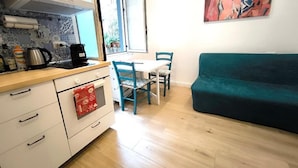 A compact kitchen with a stove, coffee maker, and a window. There is a teal sofa and a table with chairs in the living area. - Agorà Rooms (Naples)