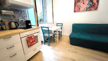 A compact kitchen with a stove, coffee maker, and a window. There is a teal sofa and a table with chairs in the living area.